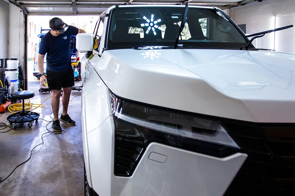 A white SUV being detailed in a garage. A man in a blue shirt and black shorts stands by the driver's side door, while bright lights reflect on the hood.
