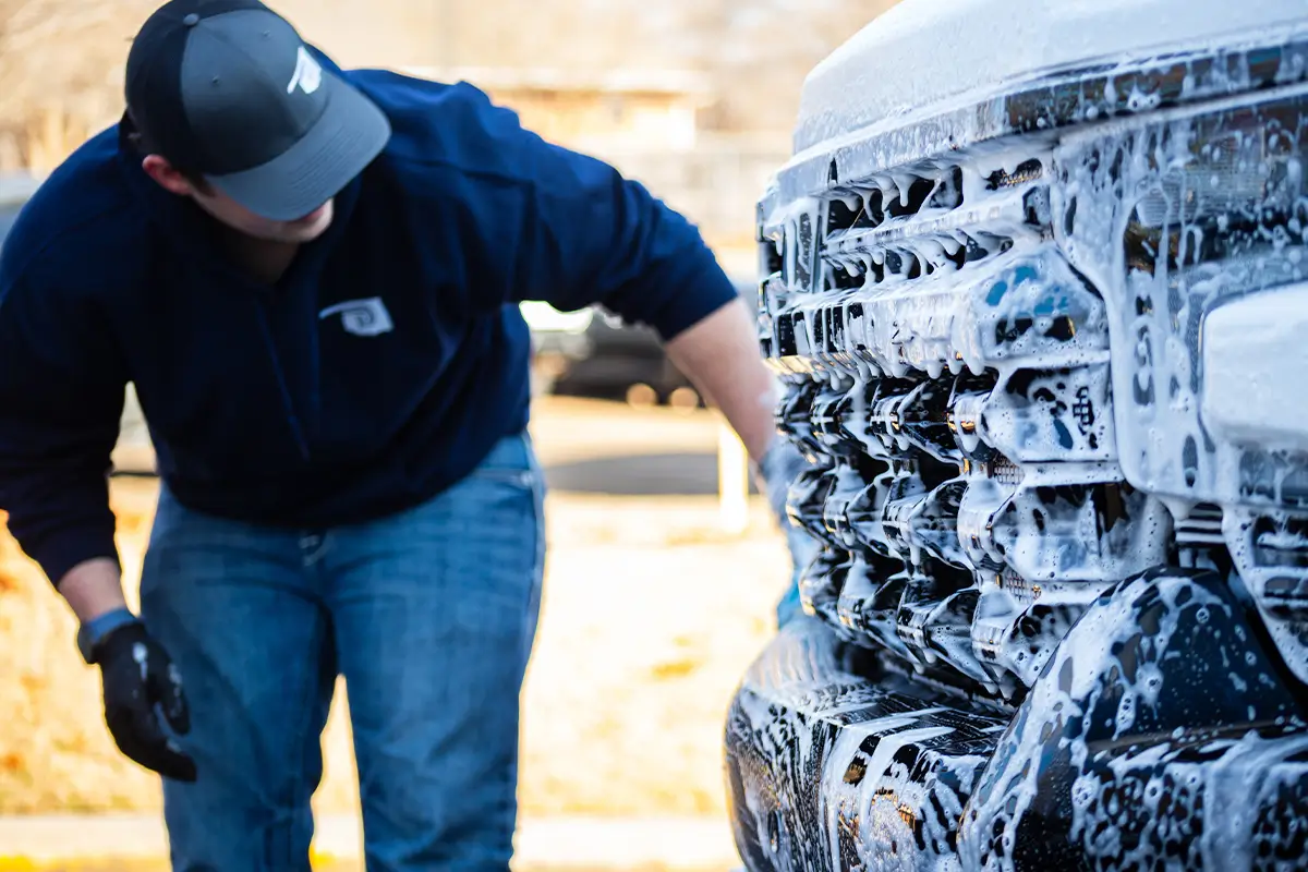 A person wearing a dark blue sweatshirt, jeans, and a baseball cap leans over to wash the soapy front grille of a white truck.