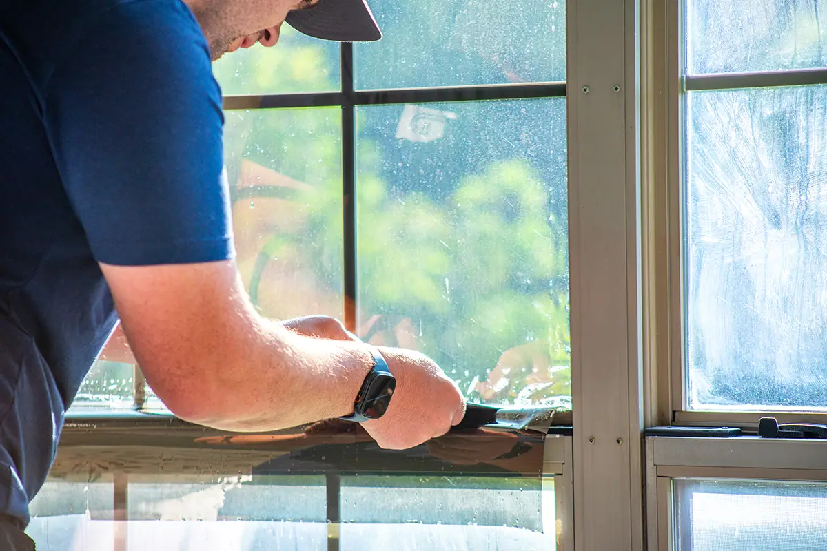 Person applying a dark solar control window tint film onto a glass window pane.