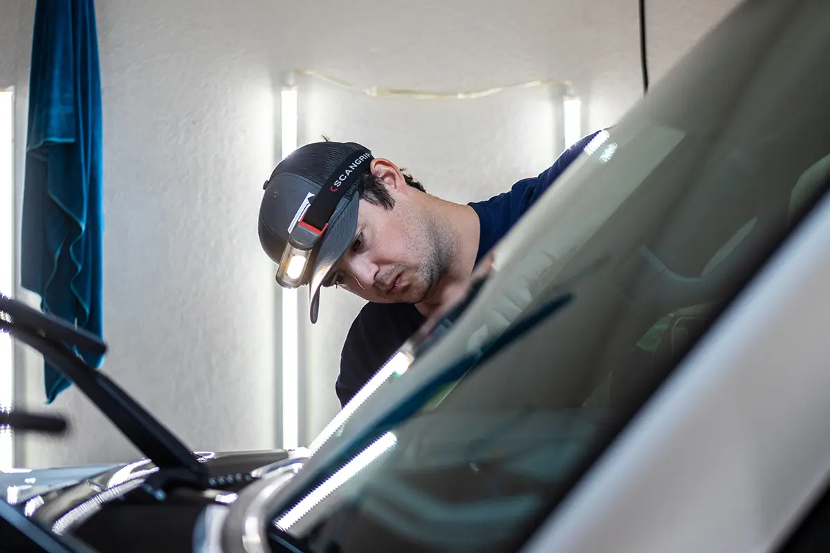 An automotive detailer wearing a Scangrip LED headlamp inspects a car windshield under bright workshop lights.